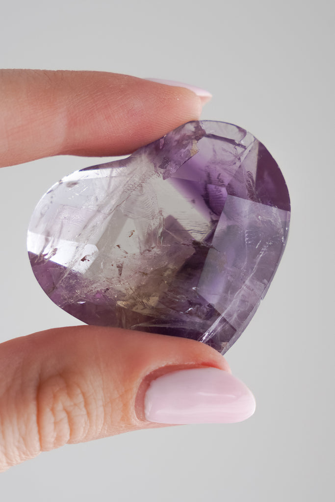 Heart-shaped amethyst crystal held between fingers against a neutral background