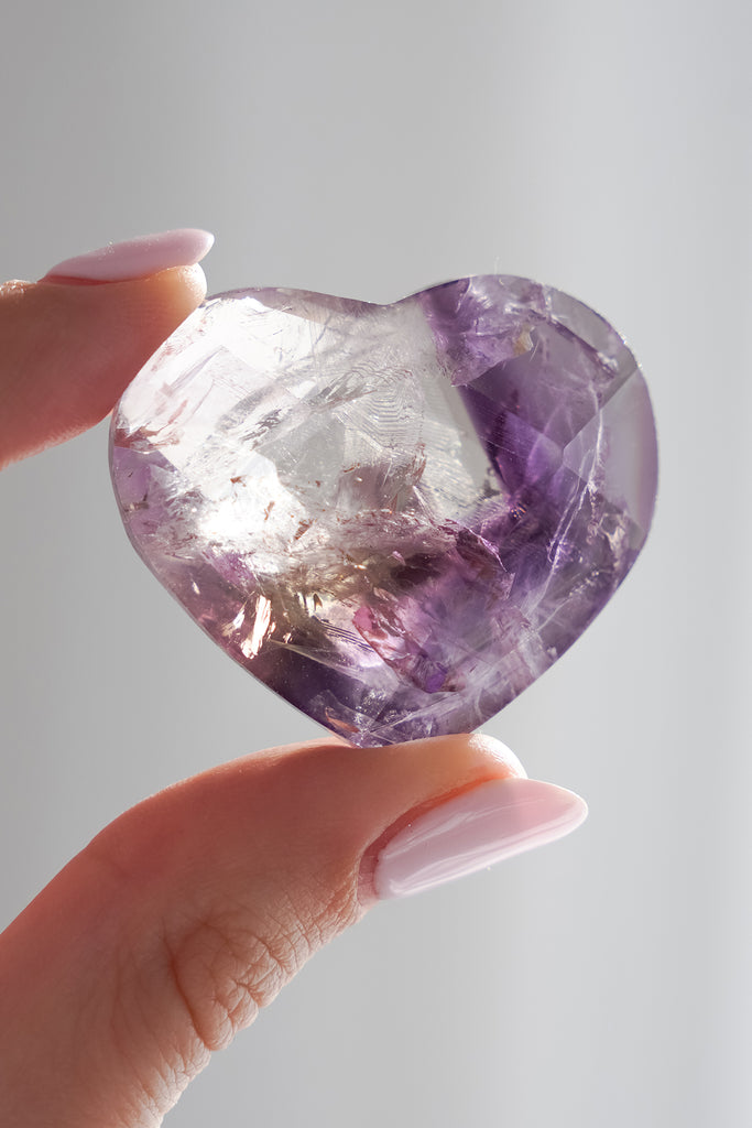 Heart-shaped amethyst crystal held by a hand with pink nail polish against a gray background