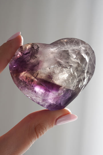 Heart-shaped amethyst crystal held by a hand against a neutral background