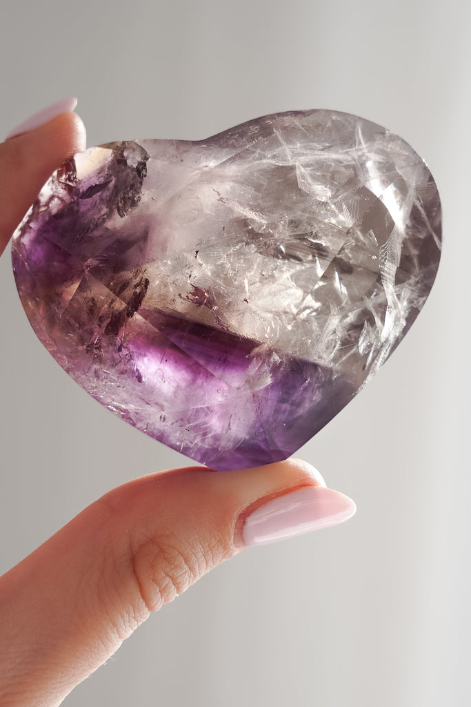 Heart-shaped purple and clear crystal held by a hand against a light gray background
