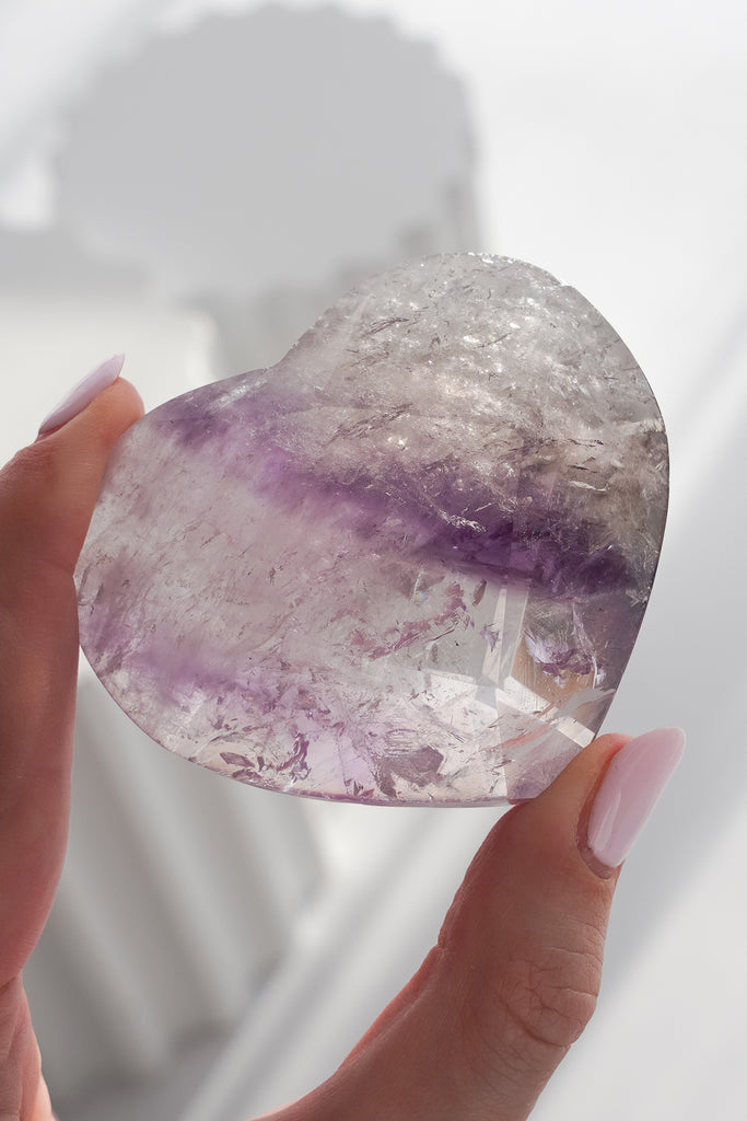 Heart-shaped crystal with purple and white colors held by a hand against a light background