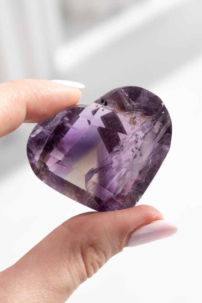 Heart-shaped purple crystal held by a hand against a neutral background