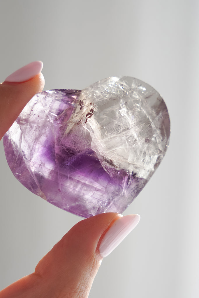 Heart-shaped amethyst crystal held by a hand against a neutral background