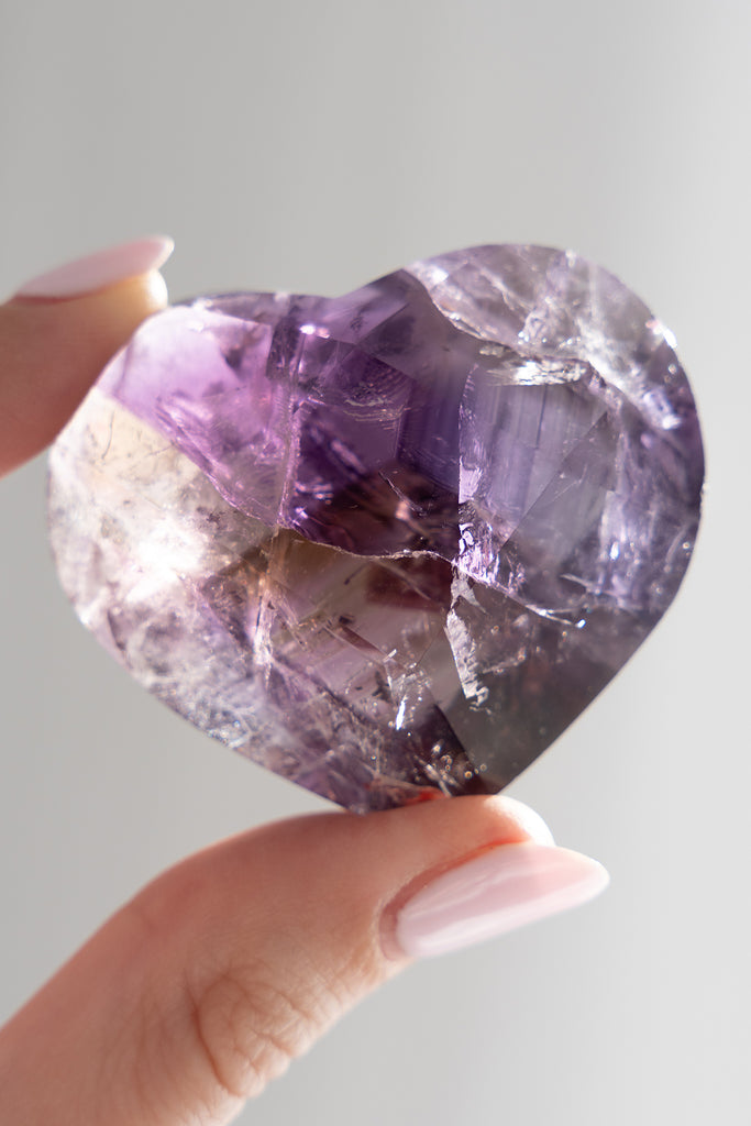 Heart-shaped purple crystal held between two fingers against a light gray background