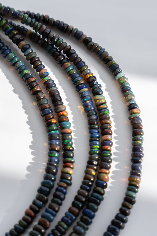 Four strands of small, dark, oval-shaped beads with iridescent opal-like flashes of green, blue, orange, and brown, arranged on a white background.