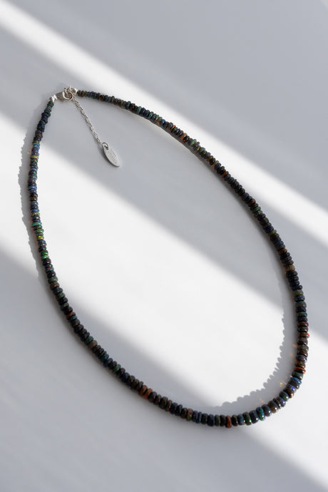 A beaded necklace made of small dark round beads with subtle iridescent green and blue tones, featuring a silver-toned lobster clasp and a small oval pendant, photographed on a white surface with diagonal shadows.