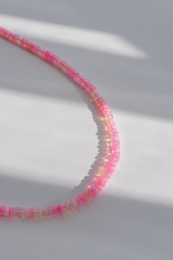 A close-up of a delicate beaded strand in bright pink and translucent tones, arranged in a curved shape on a light gray surface with soft shadows.