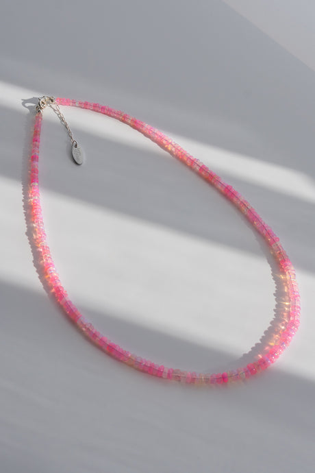 A delicate beaded necklace with alternating pink and iridescent white beads, featuring a silver-toned lobster clasp and a small oval tag, displayed on a light gray surface with soft diagonal shadows.