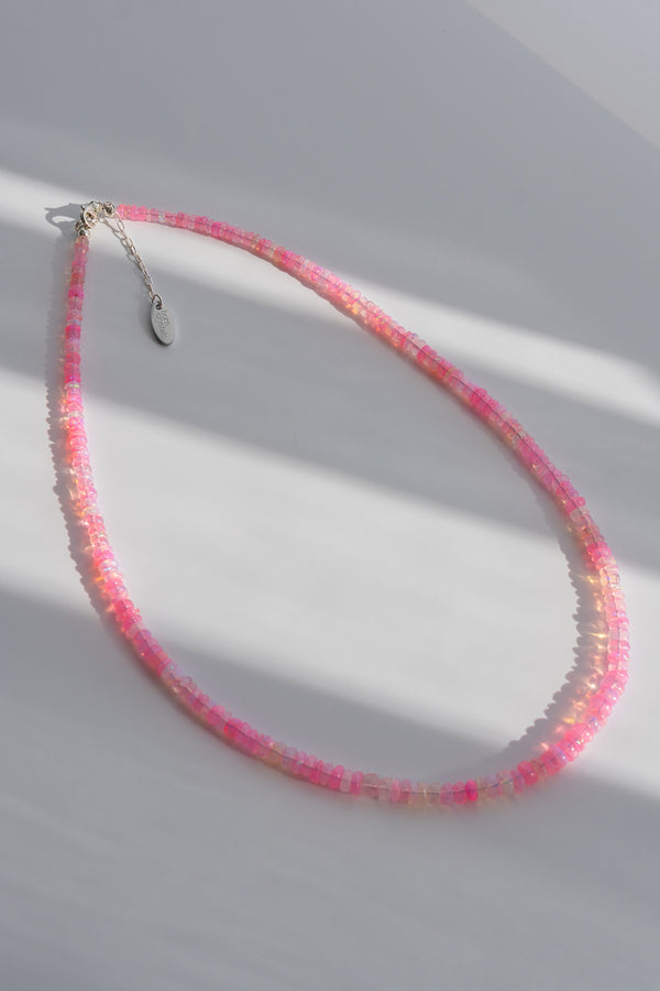 A delicate beaded necklace with alternating pink and iridescent white beads, featuring a silver-toned lobster clasp and a small oval tag, displayed on a light gray surface with soft diagonal shadows.