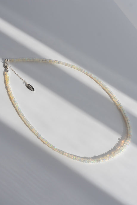 A delicate beaded necklace with small iridescent cream beads and a silver clasp, photographed on a white surface with soft diagonal shadows.